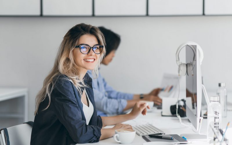 Attractive laughing freelancer girl posing with cup of coffee at her workplace. Chinese student in blue shirt works with document in campus with blonde friend in glasses.. Attractive laughing freelancer girl posing with cup of coffee at her workplace. Chinese student in blue shirt works with document in campus with blonde friend in glasses.