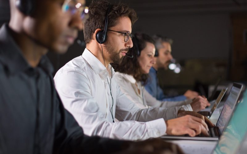 Side view of confident call center operator talking with client Side view of confident call center operator talking with client. Caucasian young man in eyeglasses typing on laptop while serving client. Call center concept