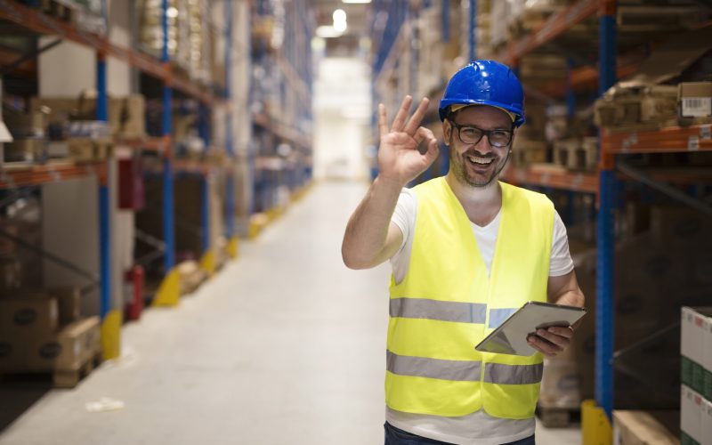 Warehouse worker standing in large storage center and showing OK hand gesture satisfied on delivering goods. Warehouse worker standing in large storage center and showing OK hand gesture satisfied on delivering goods.